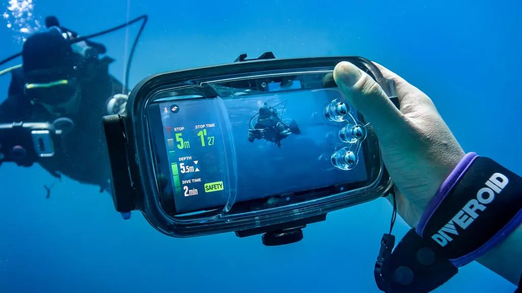 A diver underwater holding a phone in the Diveroid housing, the screen showing depth, dive time and a safety stop